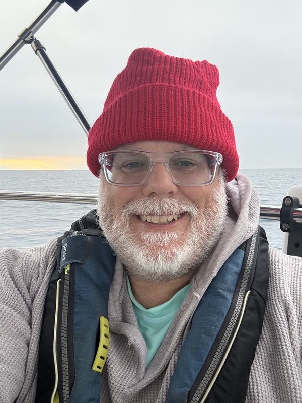 Mark A. Bunch, Sr. aboard Keeldragger in a red watch cap and inflatable PFD, open water and a low sun behind him.